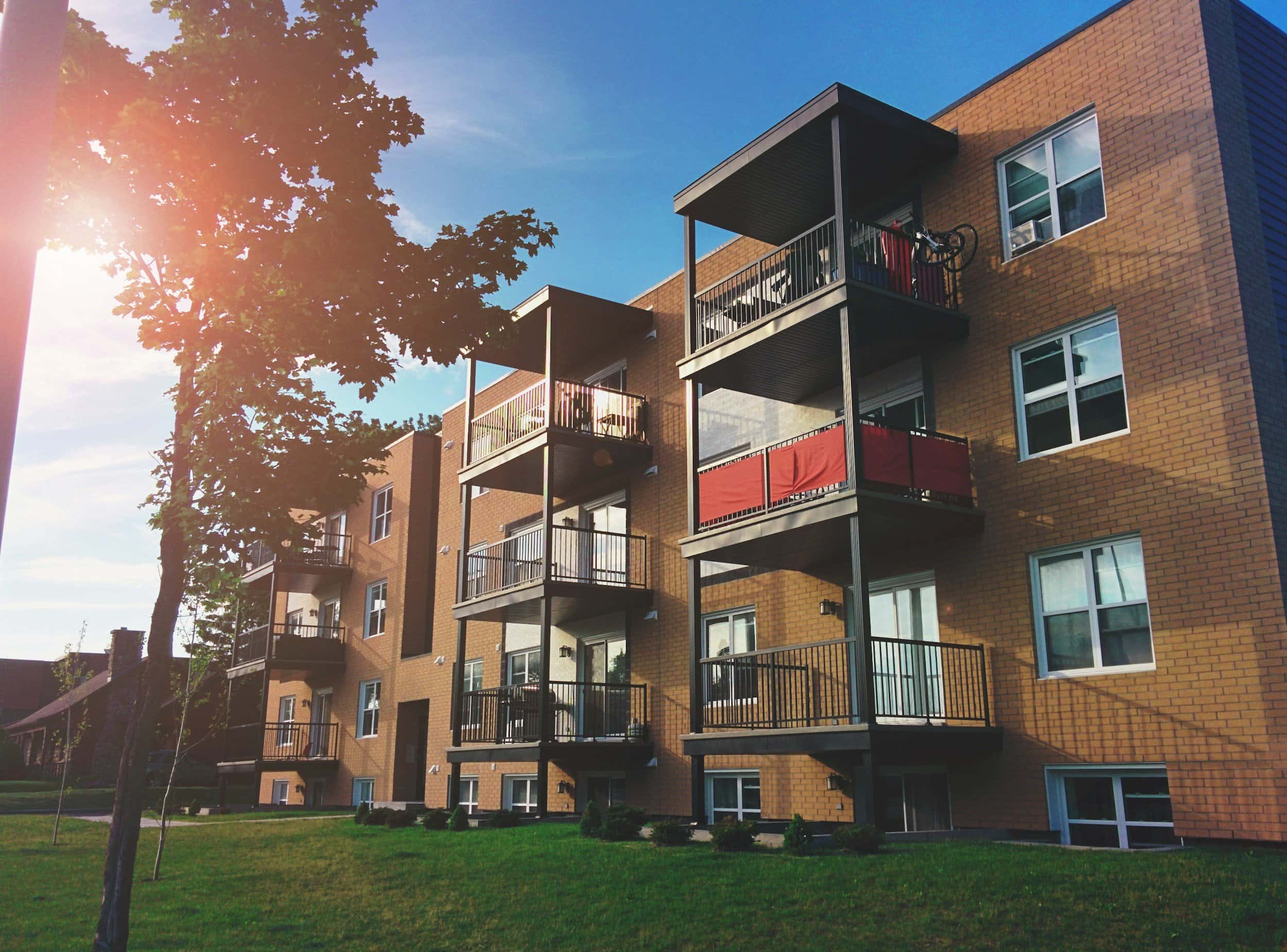 Apartment building with balconies, surrounded by greenery and trees during sunset.