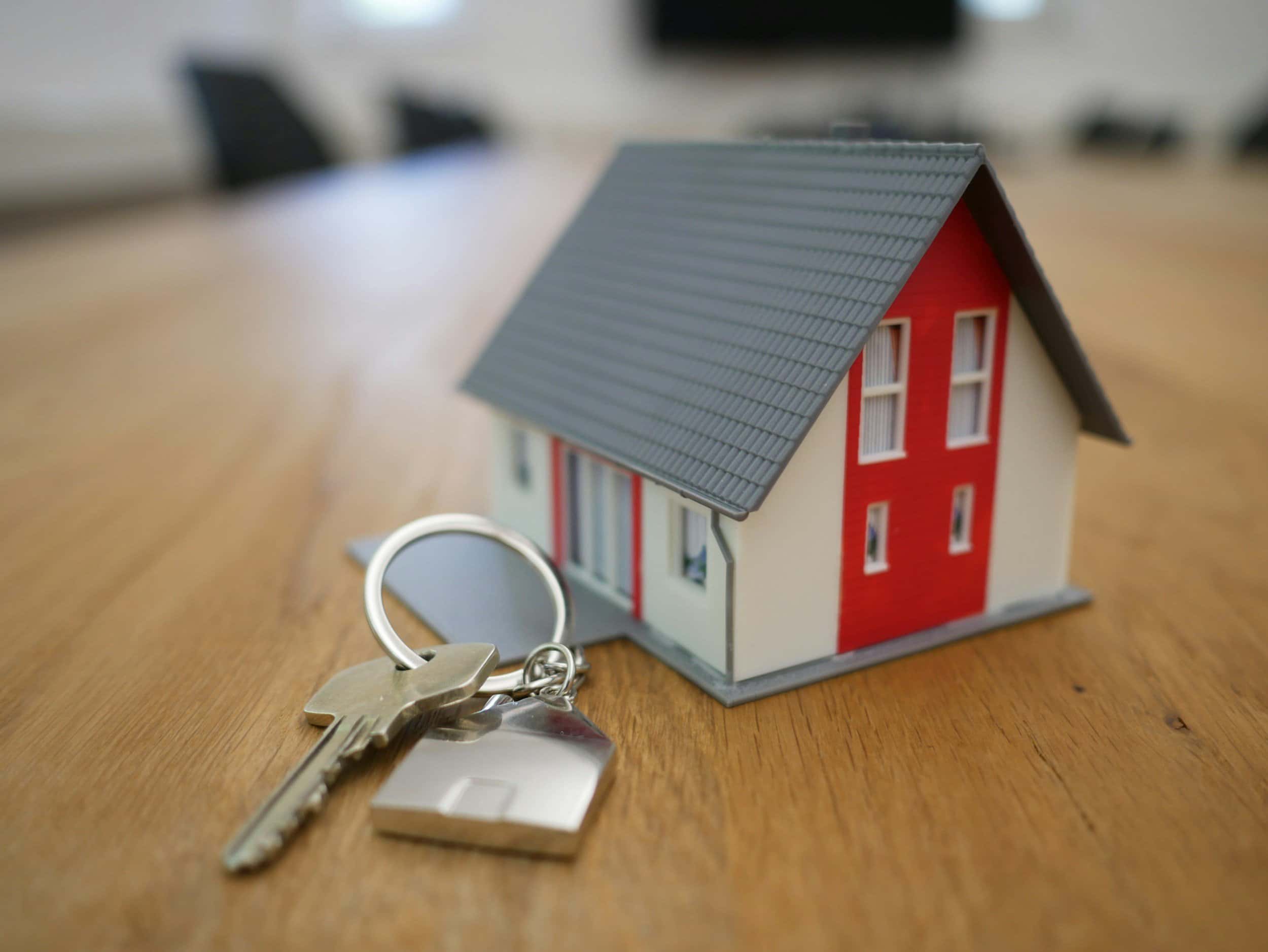 Keys on a wooden table next to a small model house with a red and white facade.